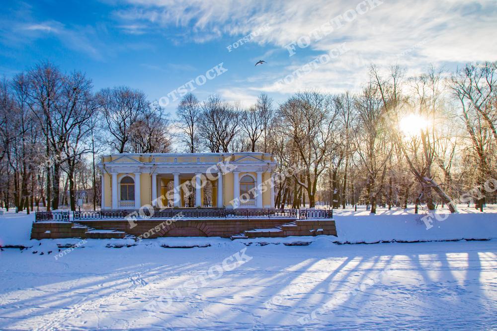 На фотографии изображён павильон Летнего сада в Санкт-Петербурге. Здание окружено заснеженными деревьями, создающими атмосферу зимнего спокойствия. Солнце пробивается сквозь ветви, добавляя тепла зимнему пейзажу. Летний сад – одно из старейших мест отдыха в городе, созданное в начале XVIII века по проекту Петра I.